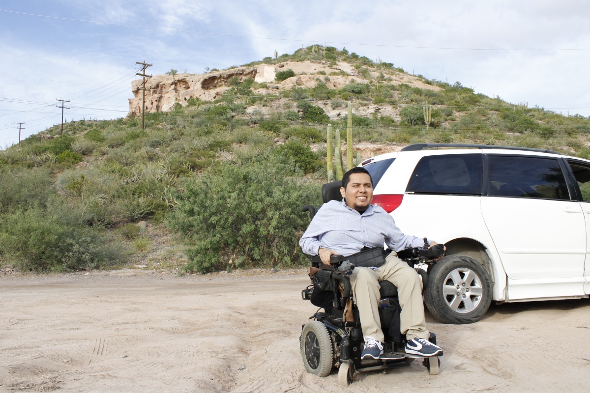 Una persona en silla de ruedas enfrente de una van y el Cerro de la Calavera en el fondo.